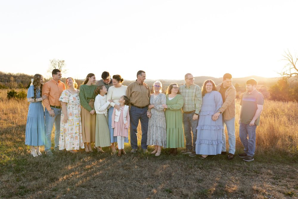 A group of people in pastel-colored dresses and formal wear pose together at sunset in a grassy outdoor setting.