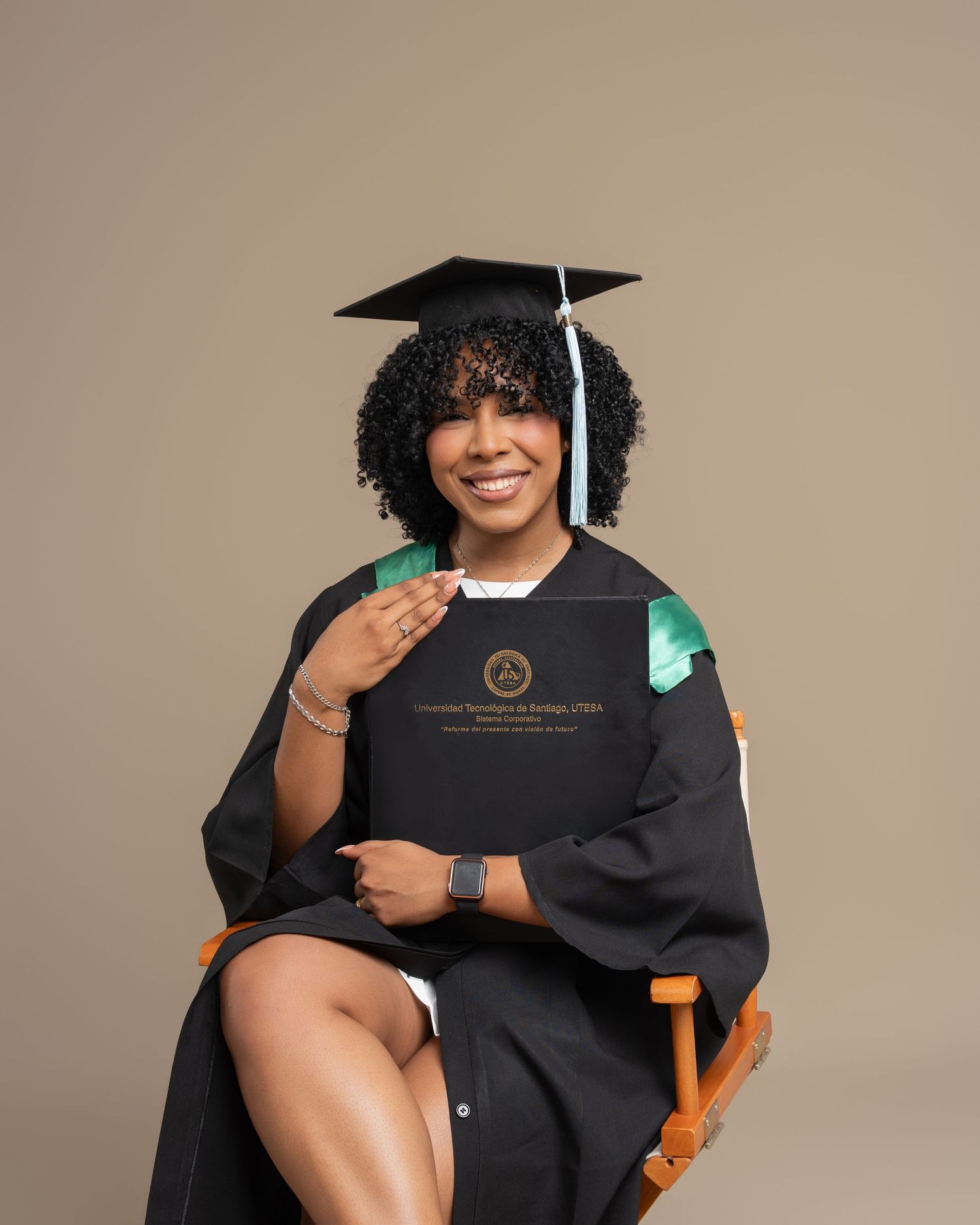 Smiling graduate in black cap and gown sits holding diploma folder, celebrating academic achievement.