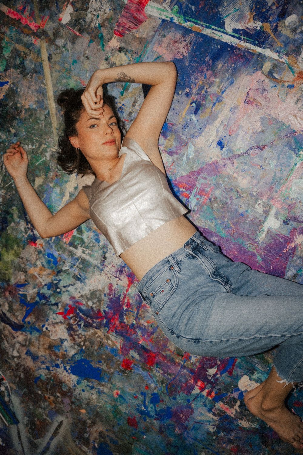 Person lying on colorful paint-splattered surface wearing denim and white top in artistic photoshoot.