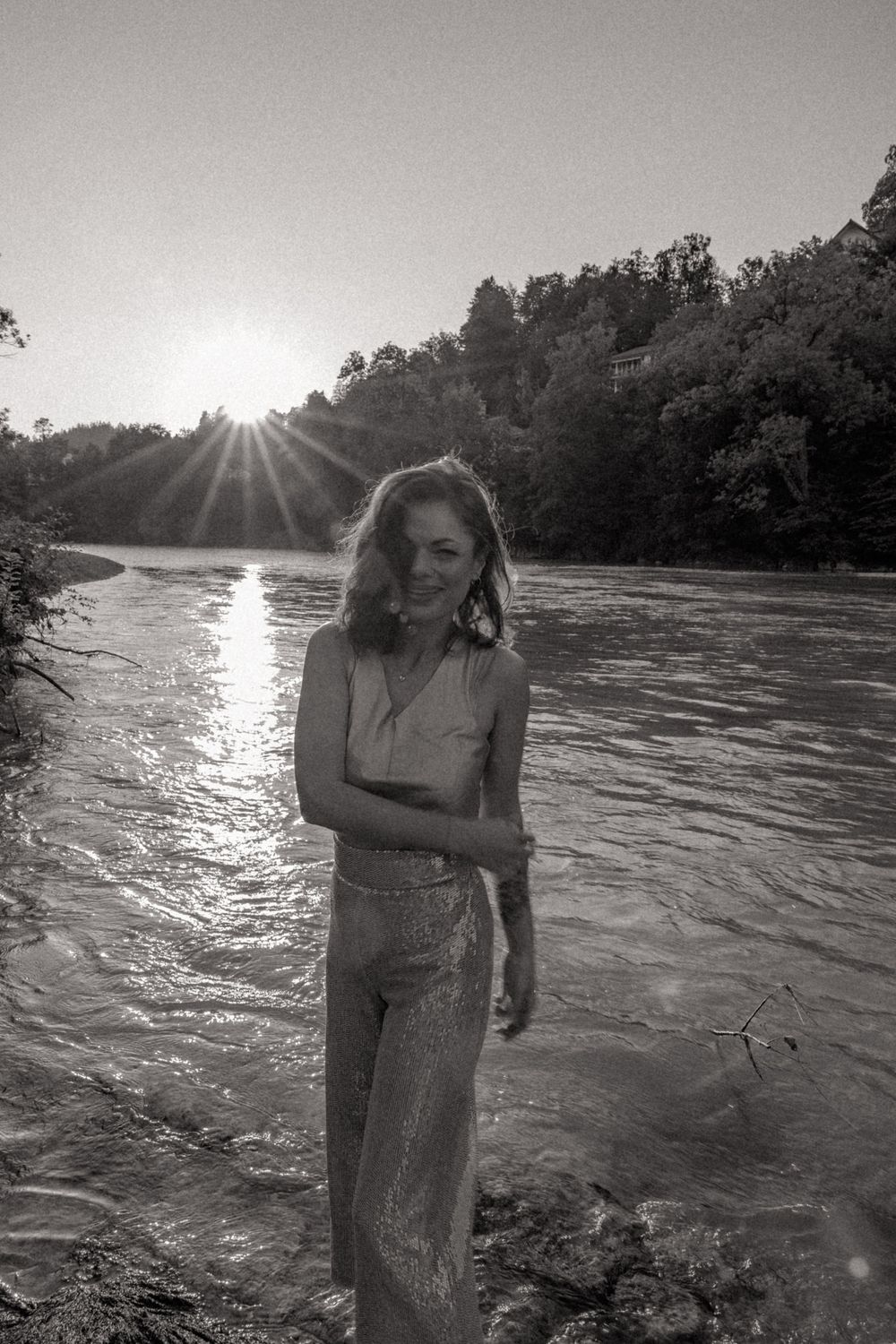 A black and white portrait by a rushing river with sun rays piercing through trees.