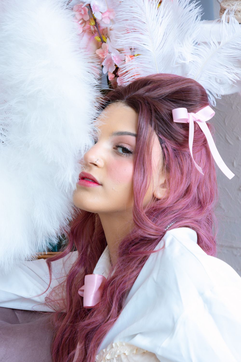 Close-up of model with pink wavy hair and bow, resting against white feather boa with floral headdress in soft light.