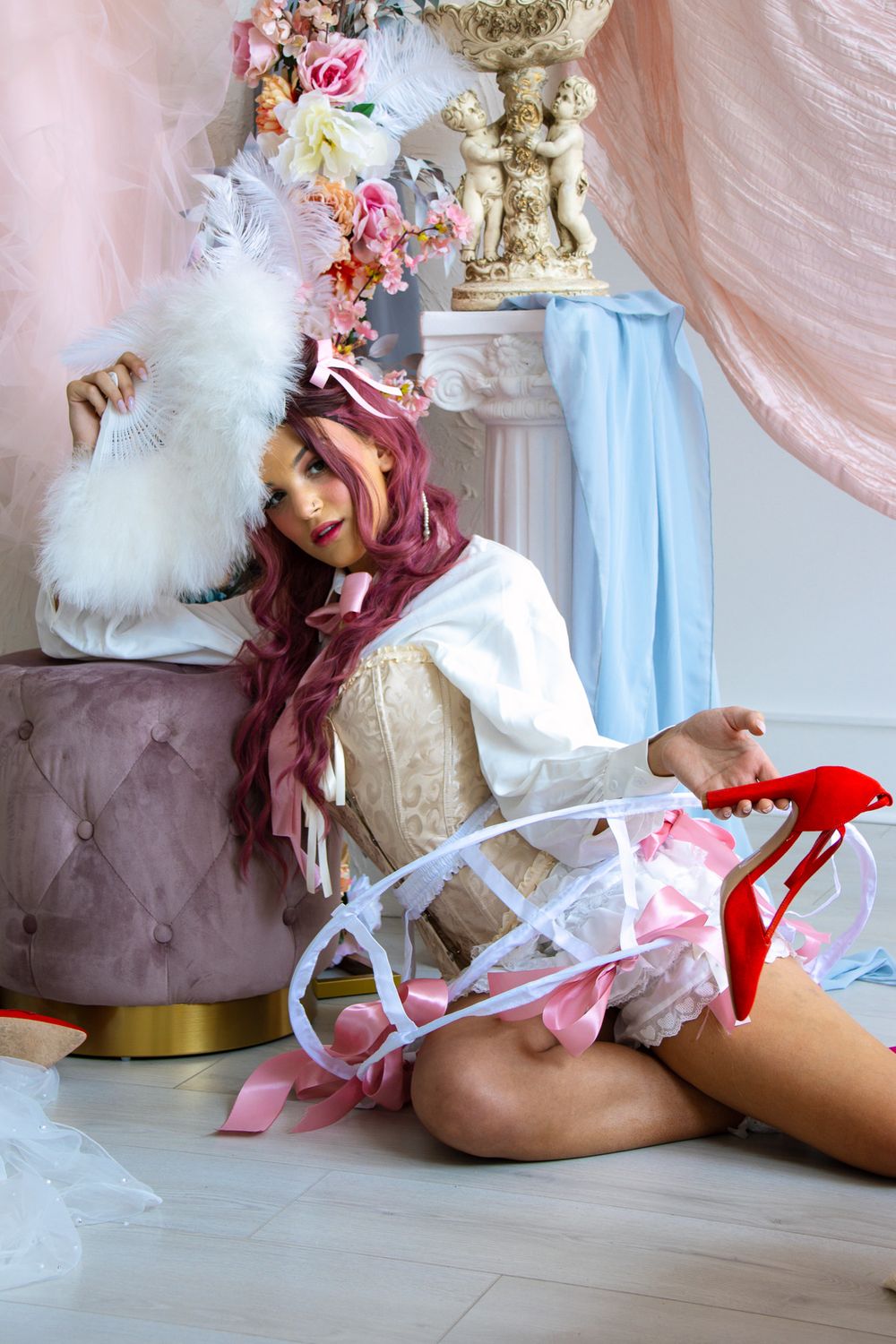 Pink-haired model in rococo costume holding red heel and feather fan, seated on floor near cherub statue and pink drapes.