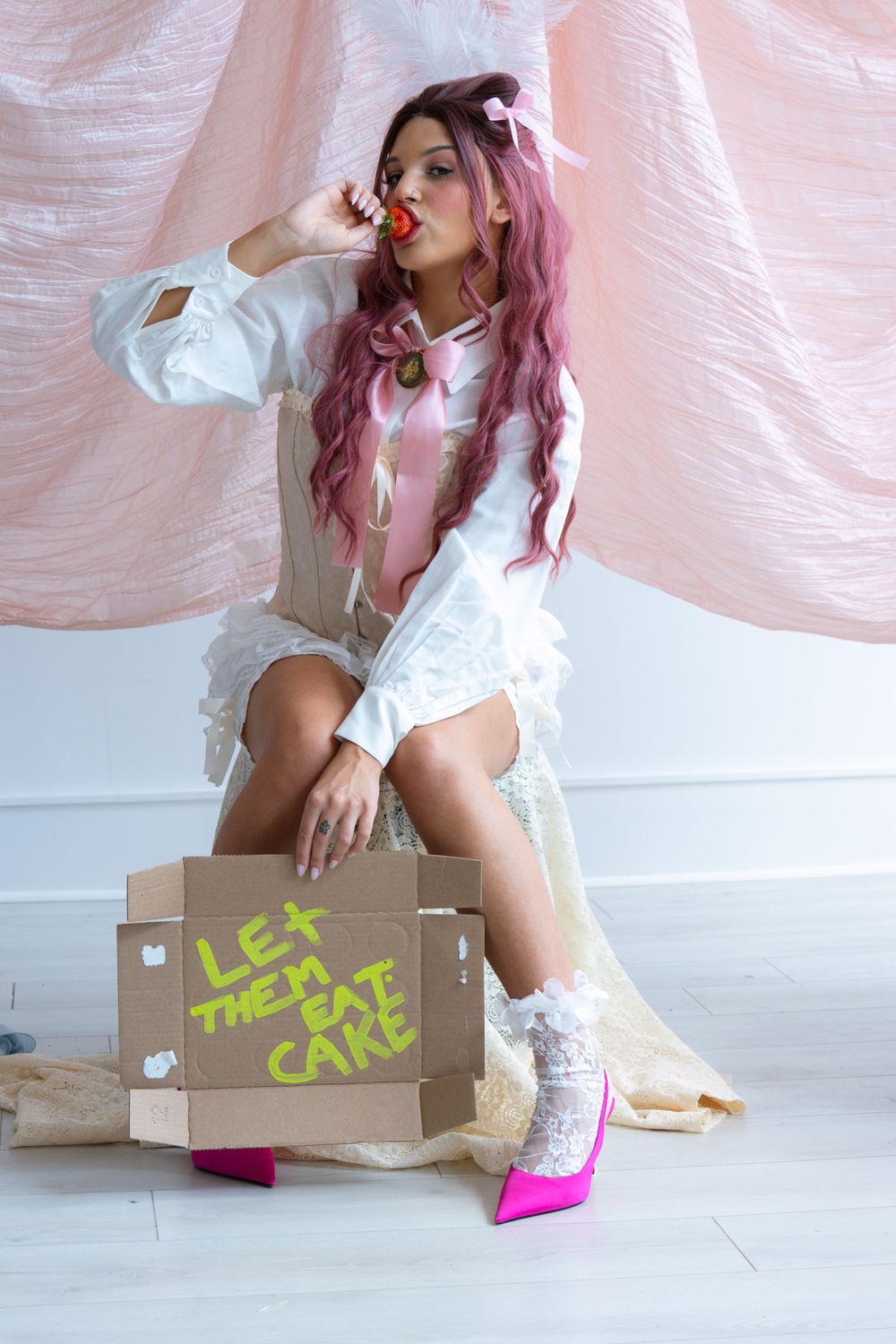 Pink-haired model in white ruffled dress eating strawberry, seated on cardboard box reading Let Them Eat Cake.