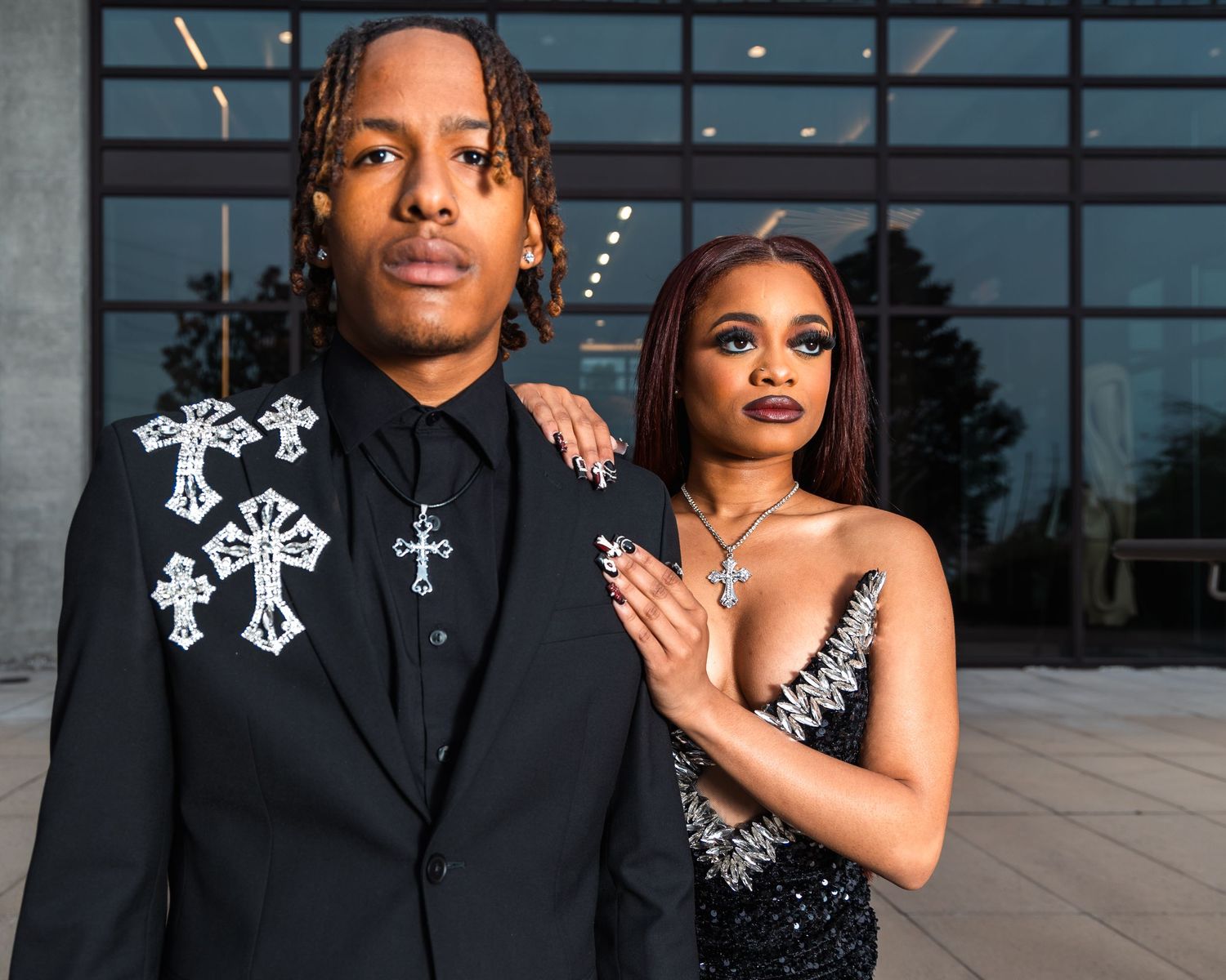 Stylish couple in formal black outfits pose outside modern building at night.