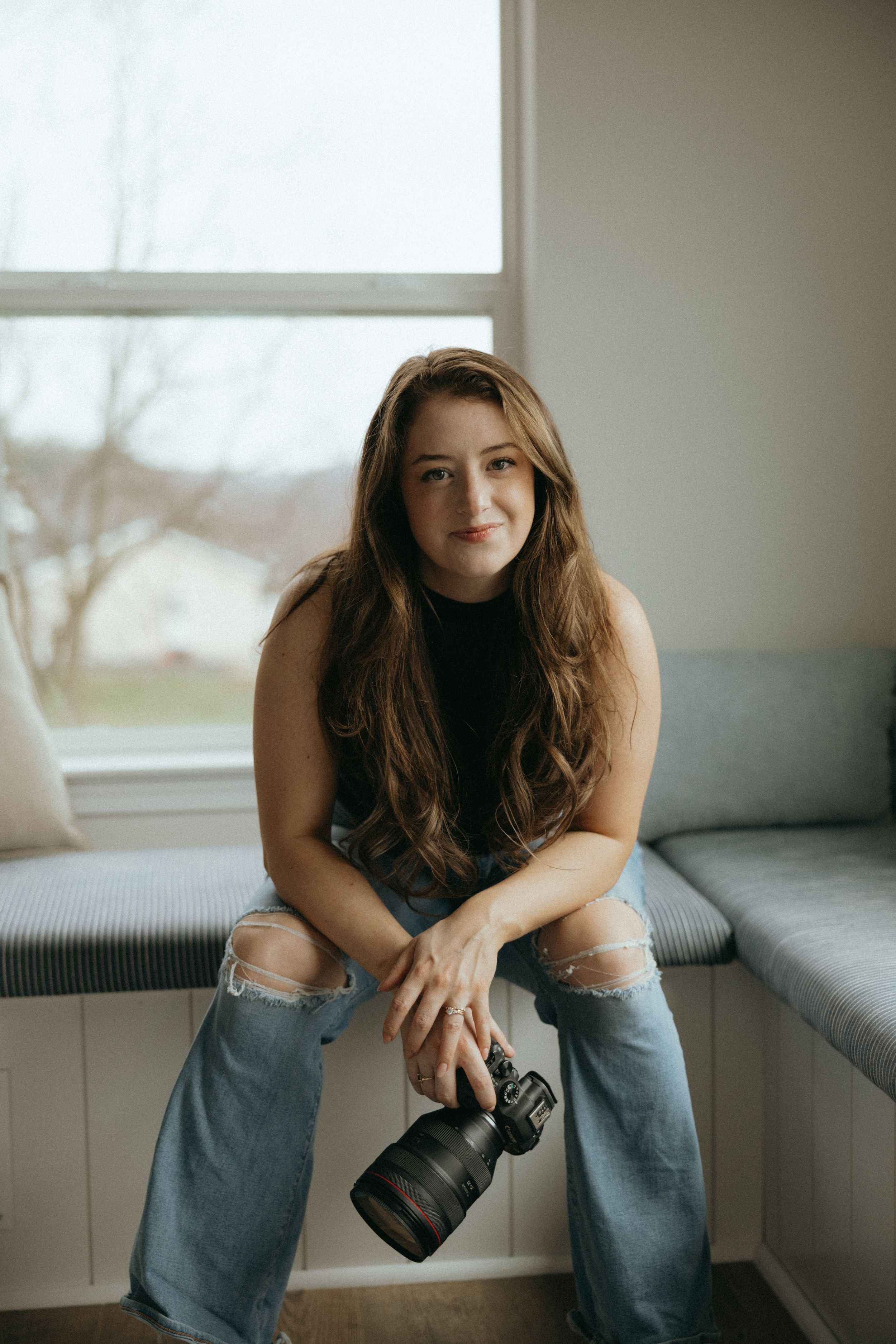 Young woman with long brown hair sits on a window bench holding a DSLR camera, wearing jeans and a black top.