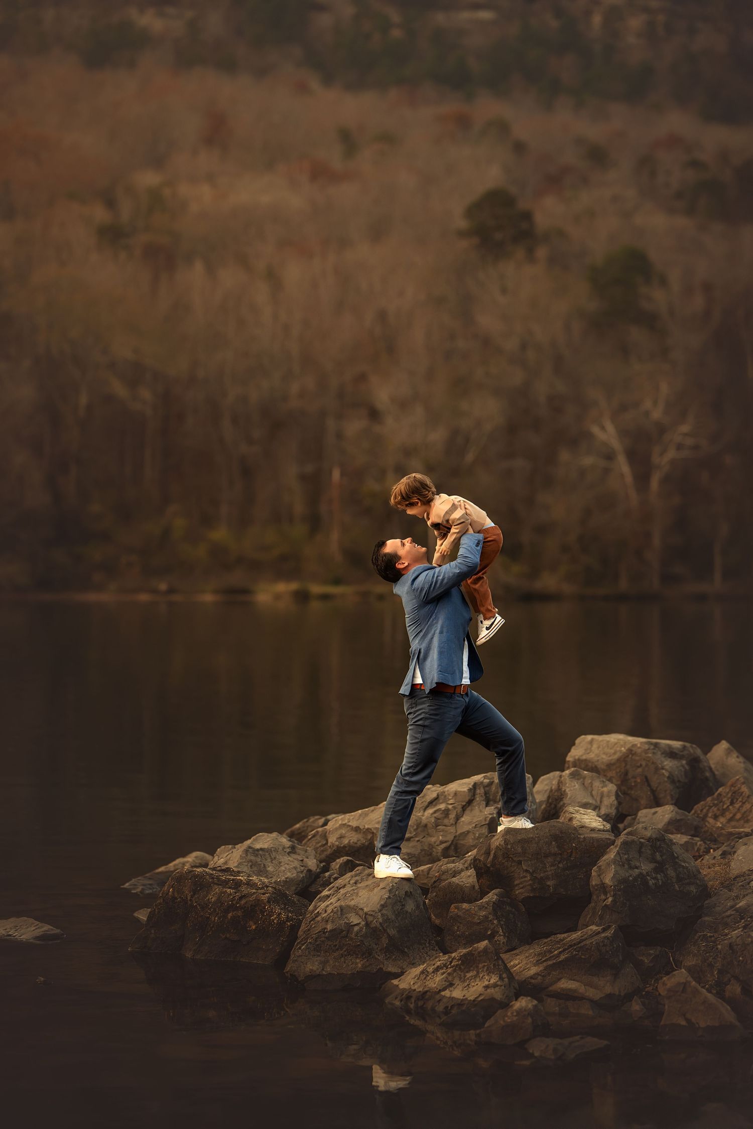 Father in blue stands on rocky shore by a still water and holds his young son up in the air for a candid playful photo.