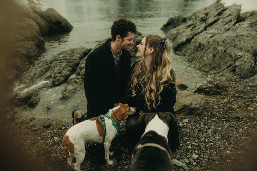 Couple sitting by rocky coastline with their dogs in moody winter setting.