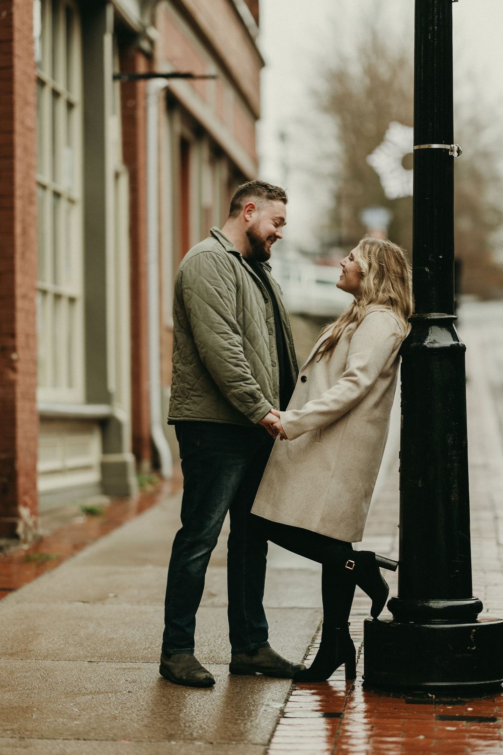 A romantic couple shares an intimate moment on a rainy city sidewalk next to a vintage lamppost.