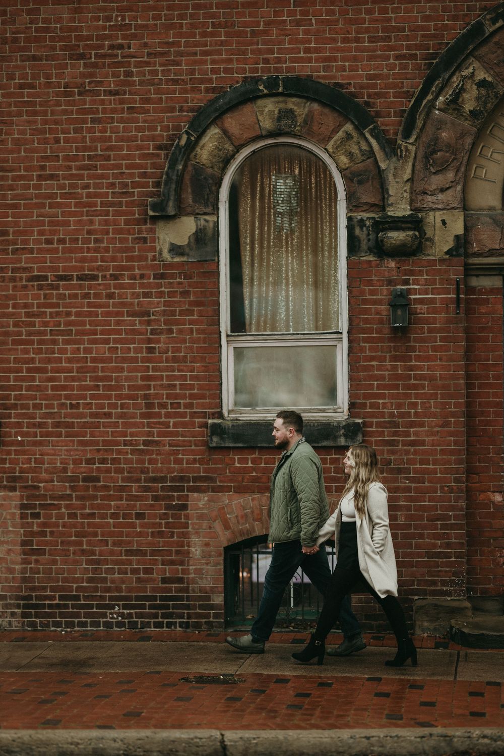 A couple walking past historic brick architecture with an arched window and ornate details.