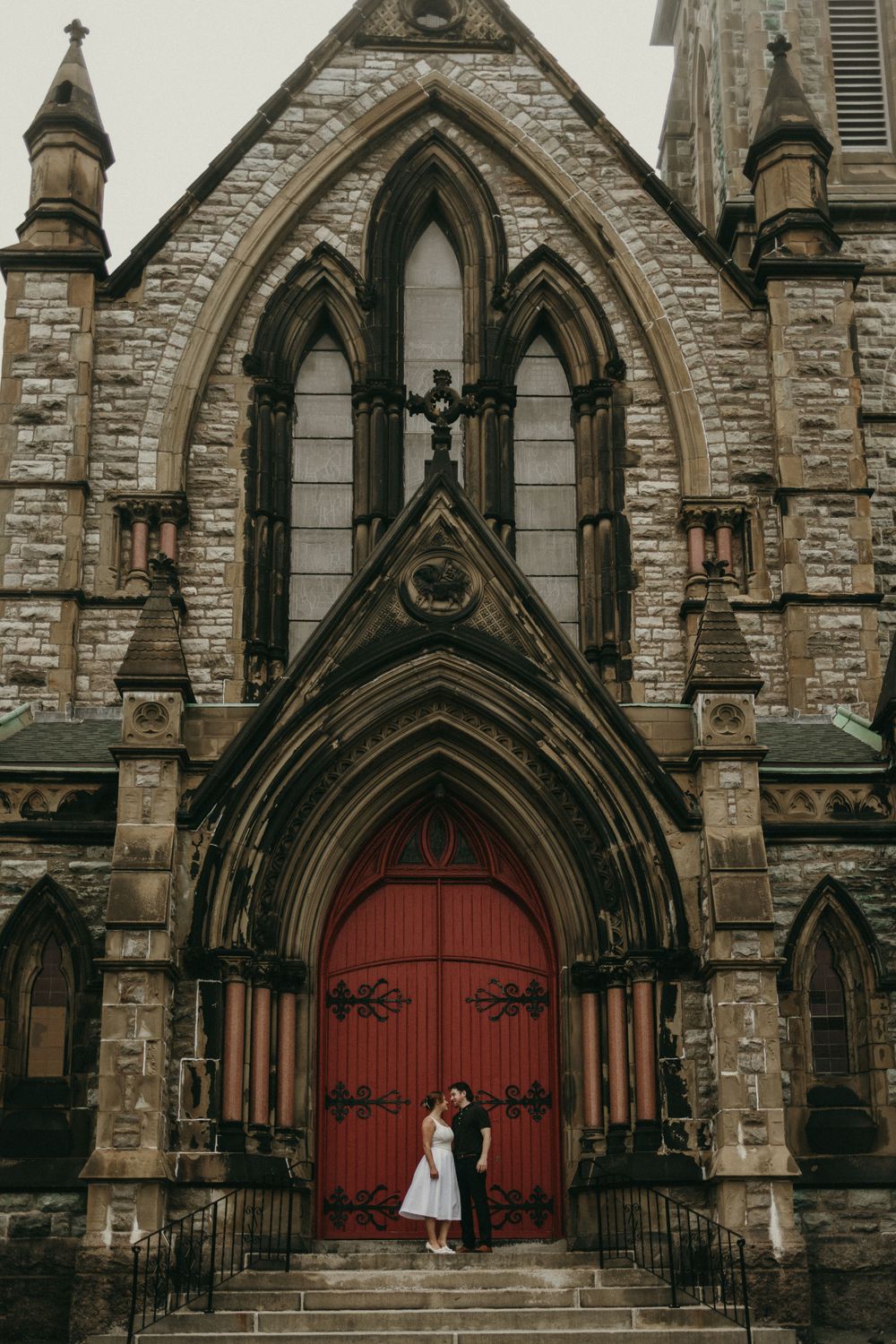 A white dress contrasts against red church doors of a Gothic stone cathedral.