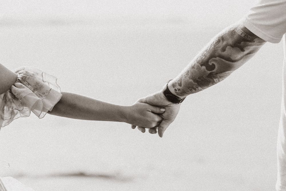 Close-up black and white shot of two hands holding each other with tattoo visible.