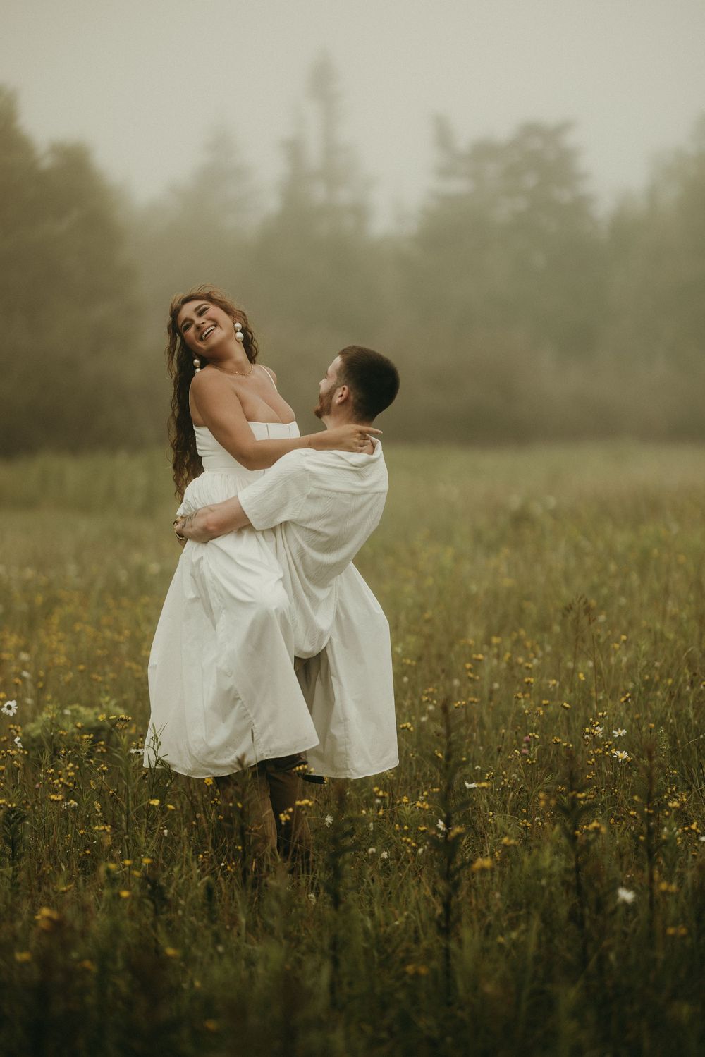 A romantic couple in white outfits embrace and twirl in a misty wildflower meadow at sunset.
