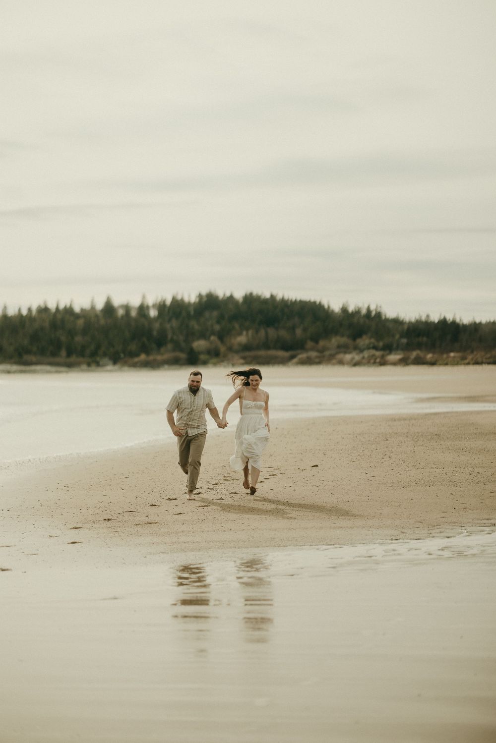 A couple runs playfully along an empty beach with evergreen trees in the background.