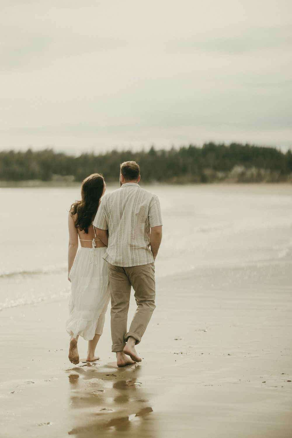 A couple walks together along the shoreline of a misty beach at sunset.
