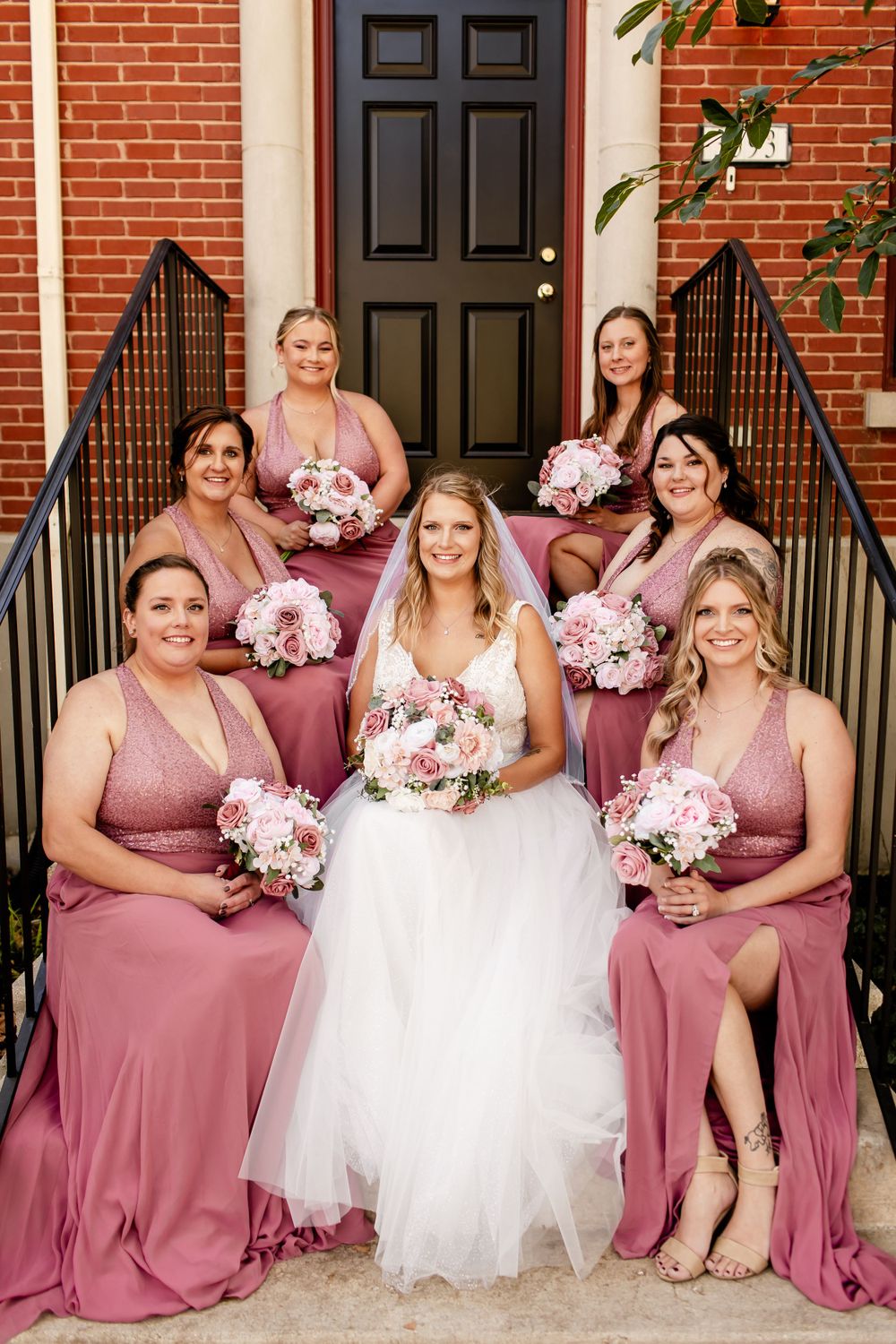 Bridal party in dusty rose dresses seated on brick church steps with floral bouquets.
