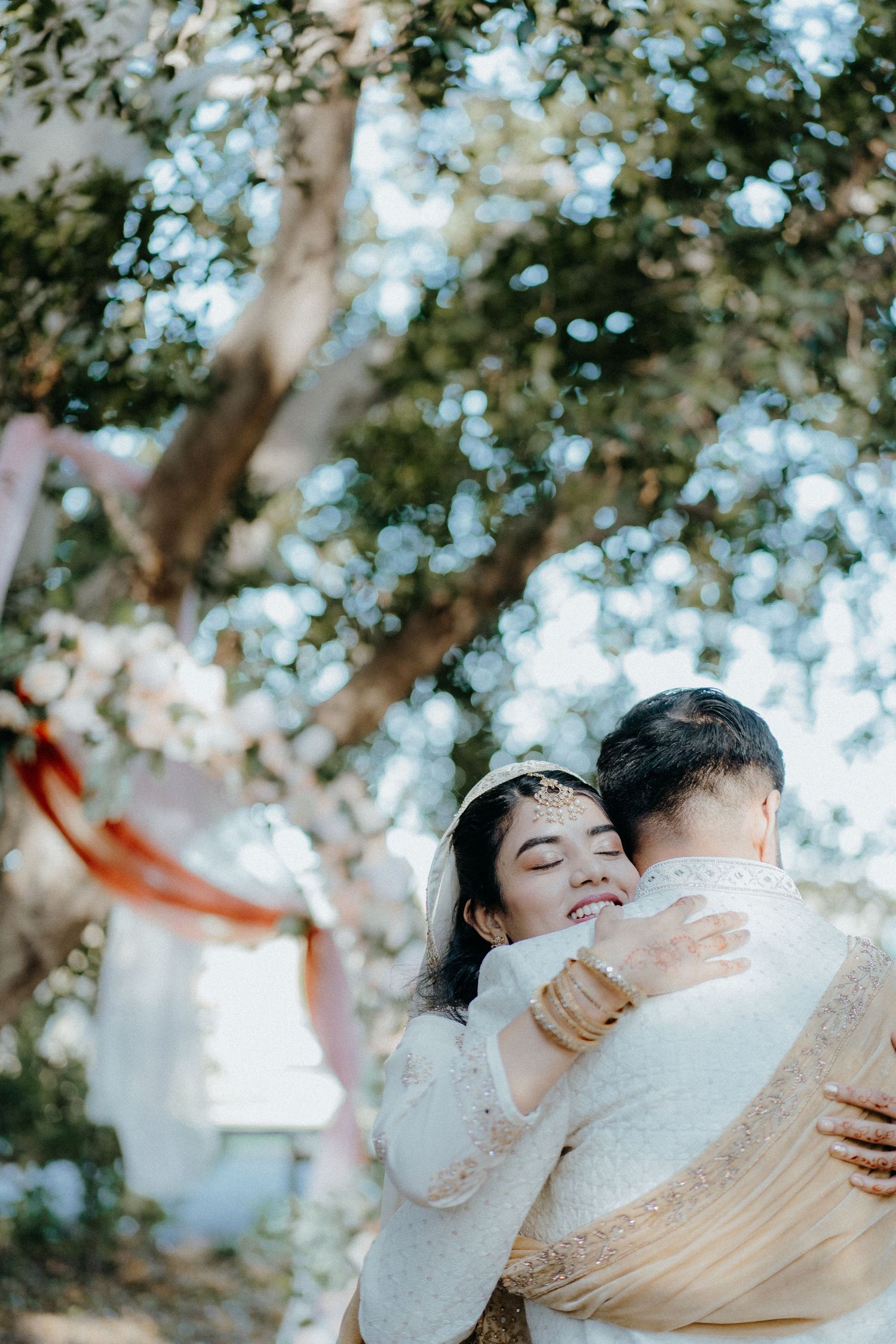 Romantic embrace under sunlit trees with white lace and flowing fabric.