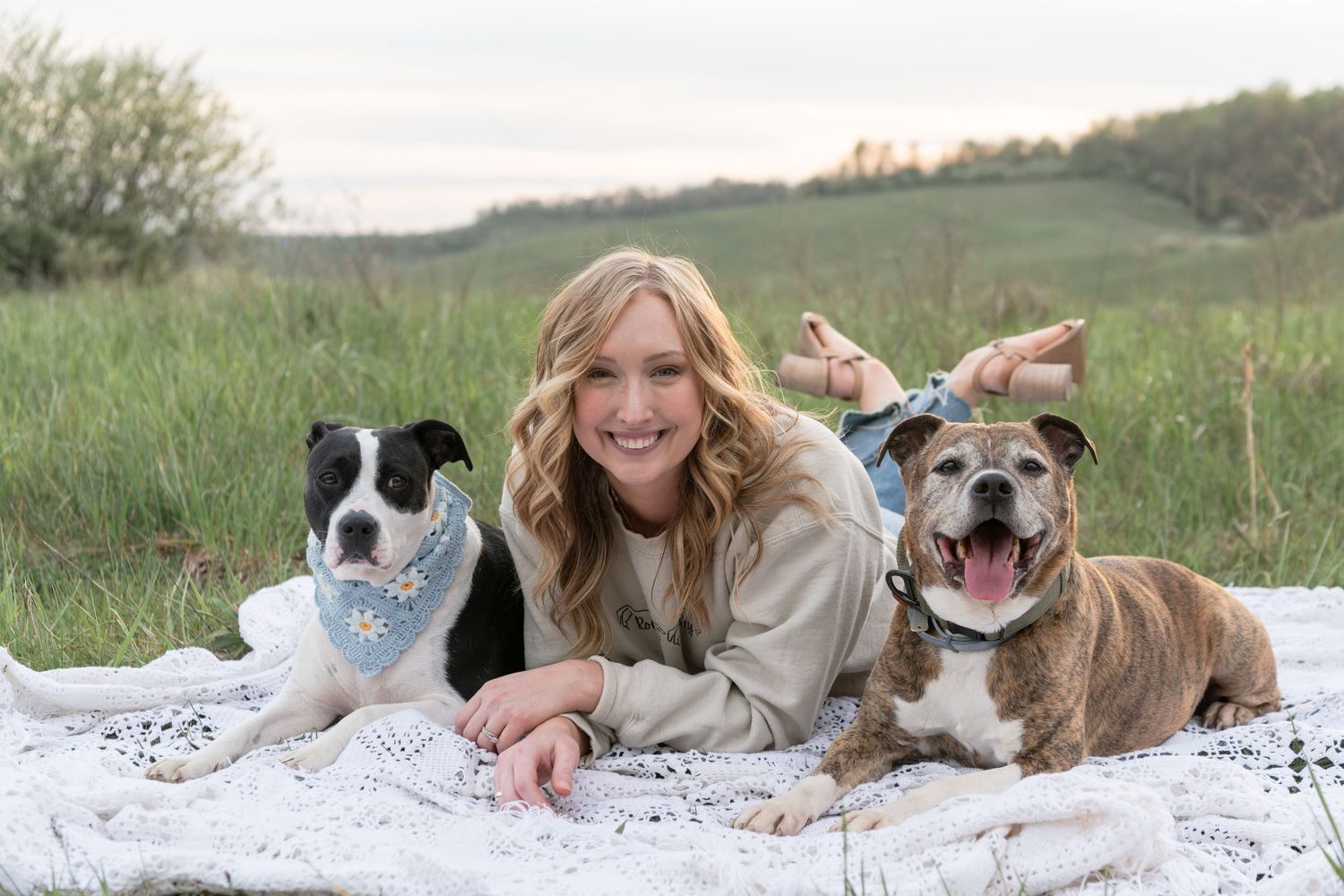 Person lying on a blanket with two happy dogs in a grassy field.