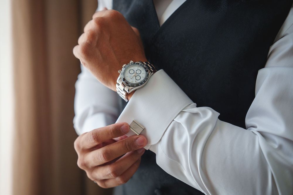 Close-up shot of hands adjusting a silver wristwatch against a white shirt sleeve and dark suit jacket.