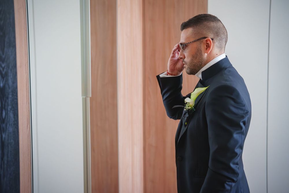 A groom in a navy suit adjusts his tie while getting ready in a room with vertical striped wallpaper.