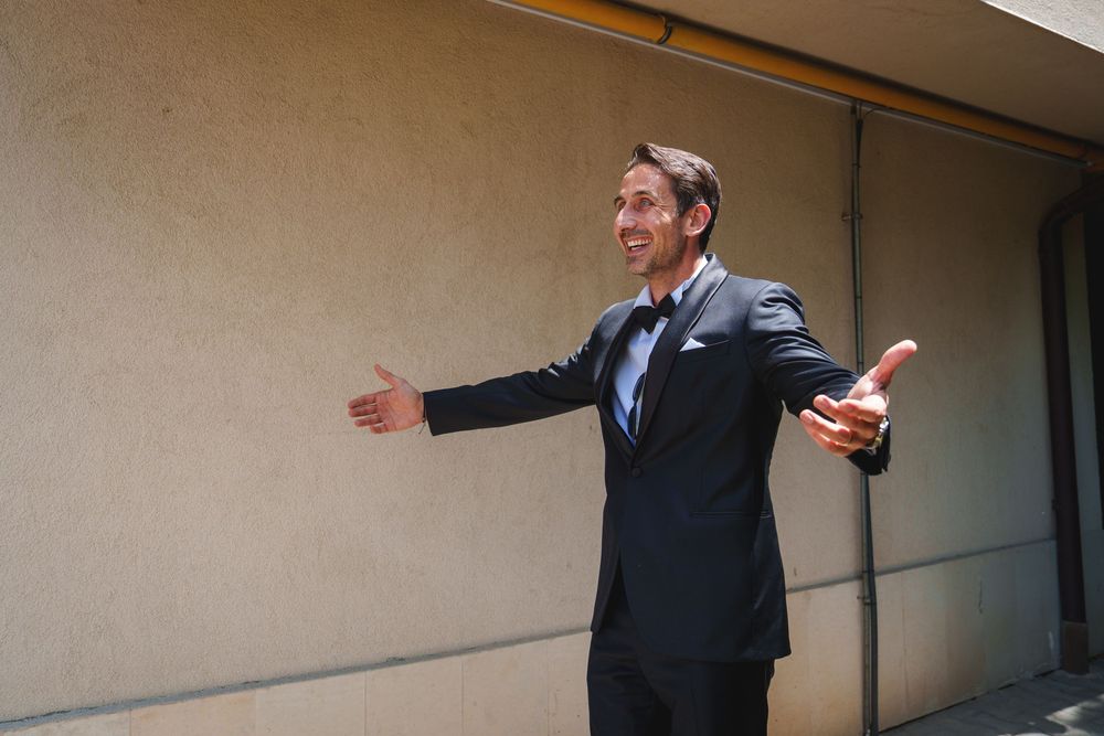 A person in a dark business suit stands with arms outstretched in a welcoming gesture against a beige wall.