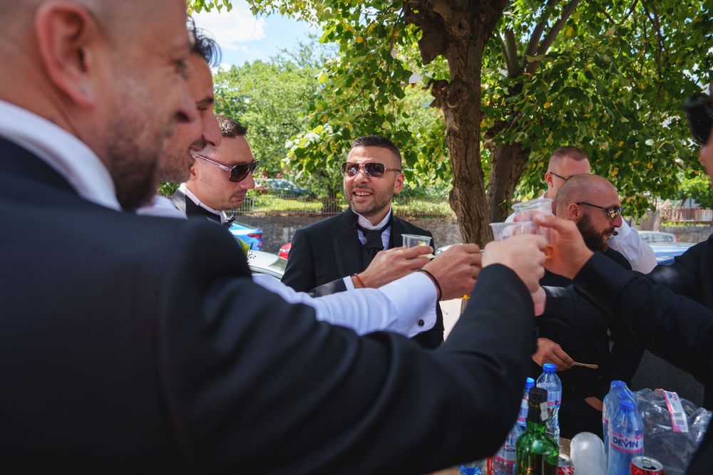 A group of formally dressed people outdoors raise their glasses in a celebratory toast under green trees.