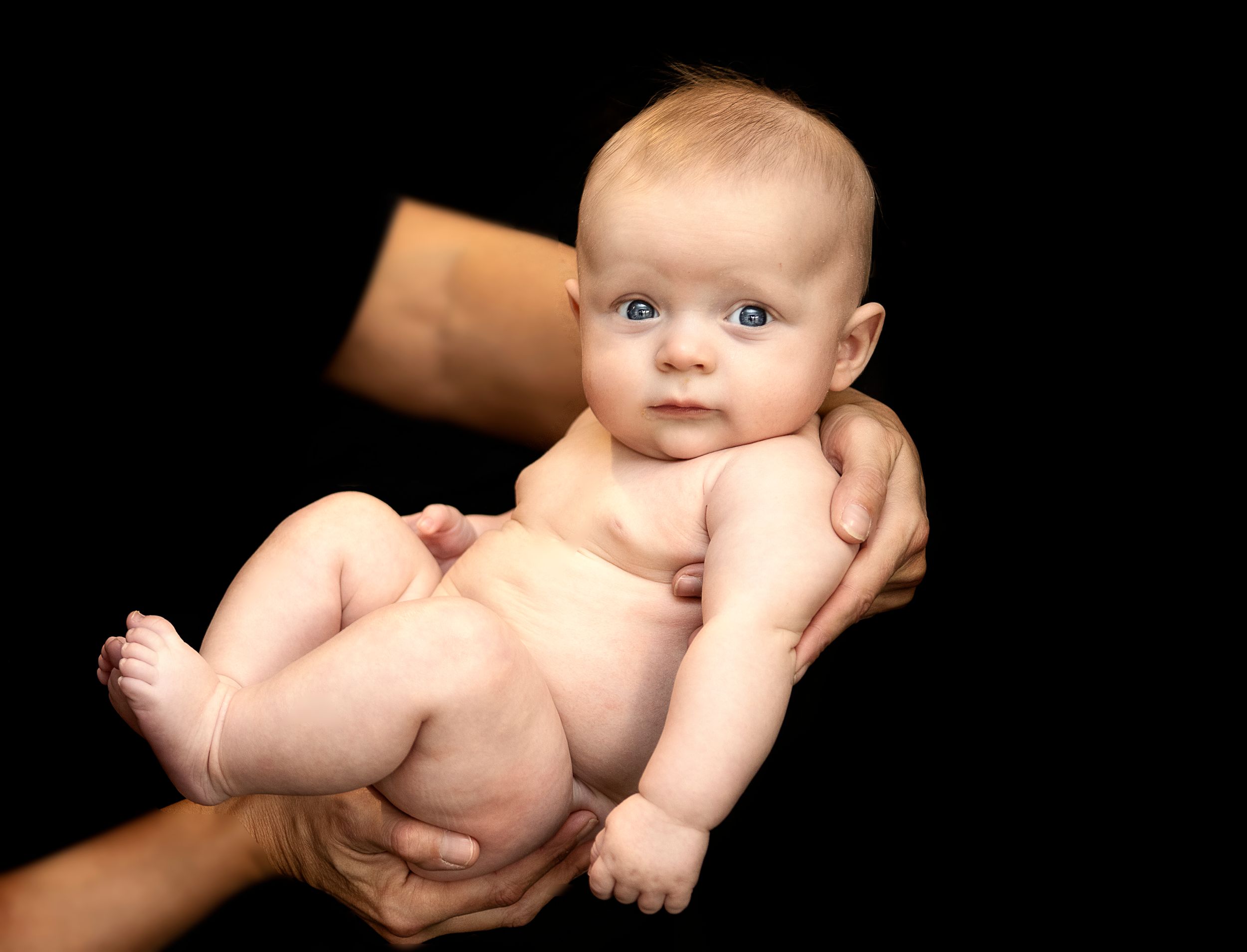 Adorable blue-eyed baby held gently in adult hands against a black background.