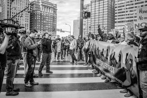 Black and white photo of urban protest with crowd holding banner, media filming, city buildings in background.
