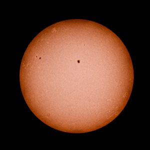 A telescopic image of the sun showing visible sunspots on its orange surface against a black background.