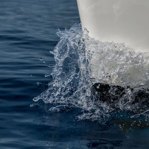 Close-up of ocean water splashing dramatically against the white hull of a sailing boat on deep blue sea.
