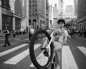 Black and white photo of a young person posing with a BMX bike on a city street with tall buildings in the background.
