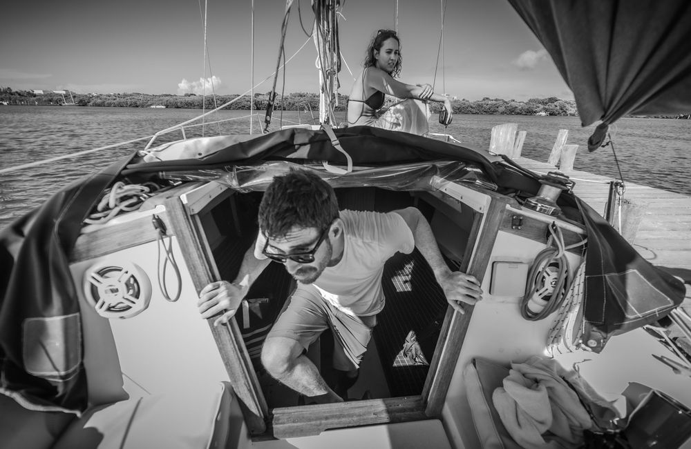 Black and white photo of two people on a sailboat, man emerging from cabin hatch on open water.