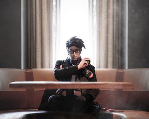 Person in black jacket and glasses with dark hair poses at a table in dramatic window lighting.