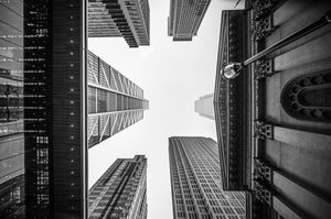 Black and white worm's eye view of towering skyscrapers converging toward a bright sky in a city.