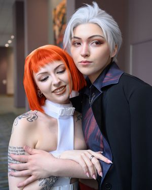 Two cosplayers pose together, one with orange bob hair in white dress, other with silver hair in black suit with red tie.