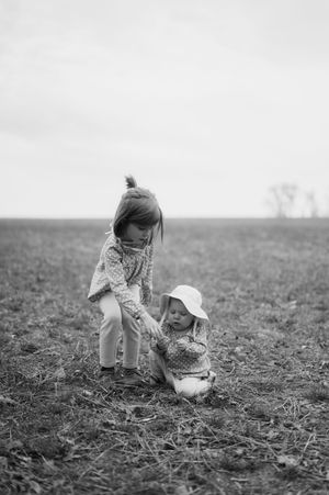 Two young children playing in an open field, older girl helping toddler in sun hat, black and white photography.