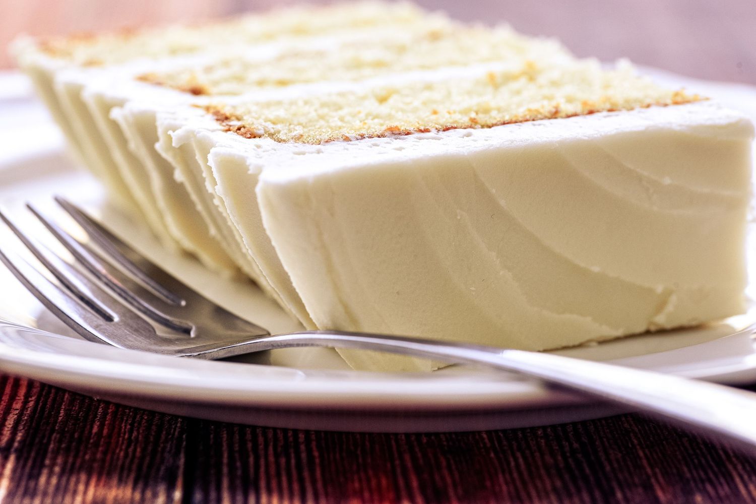 A slice of white cake with creamy vanilla frosting on a white plate with a silver fork.