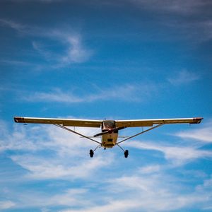 Small propeller plane flying low against a bright blue sky with wispy white clouds, viewed from below.