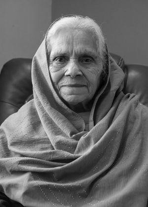 Elderly woman wrapped in a traditional shawl over her head, sitting in a chair, black and white portrait photo.