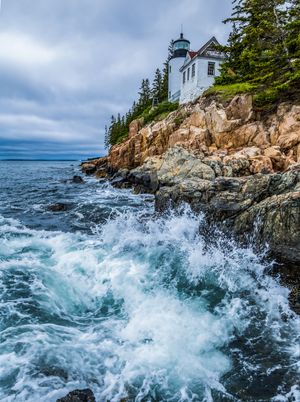 Waves crashing on rocky coastline below a white lighthouse surrounded by evergreen trees under cloudy skies.