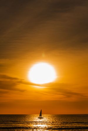 Sailboat silhouette on calm ocean waters during a stunning golden sunset with glowing sun and dramatic orange sky.