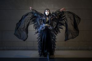 Person in dramatic black feathered costume with wings and white mask poses elegantly against a stone wall.