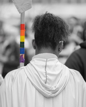 Person in white hoodie holding a rainbow Pride flag at an outdoor event, black and white photo with colorful flag.