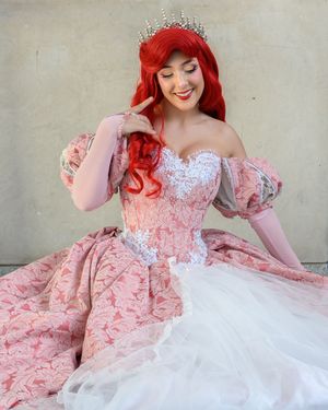 Woman in pink princess gown with red hair and tiara poses elegantly, cosplaying as Ariel from The Little Mermaid.