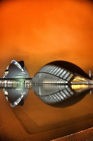 Futuristic architecture of City of Arts and Sciences in Valencia reflecting on water under orange night sky.