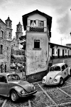 Black and white photo of two vintage VW Beetles parked on cobblestone street in historic Mexican town with church towers.