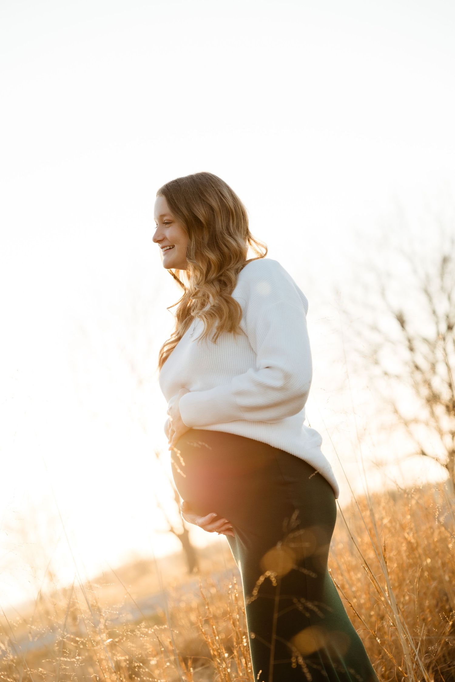 A joyful pregnant woman in a white sweater cradles her baby bump outdoors in a golden sunlit field.