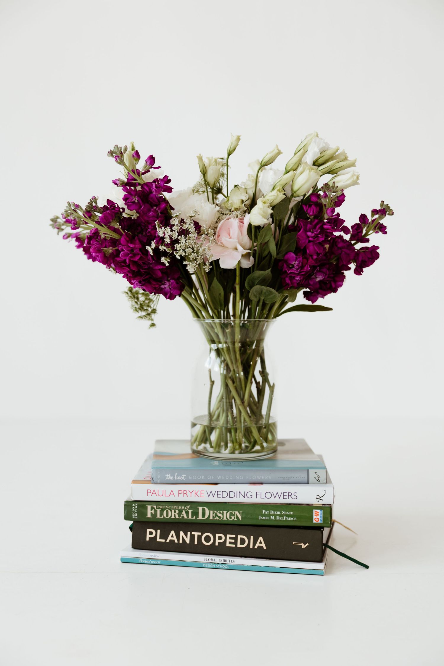 Purple and white flowers in glass vase atop stacked floral design books on white surface.