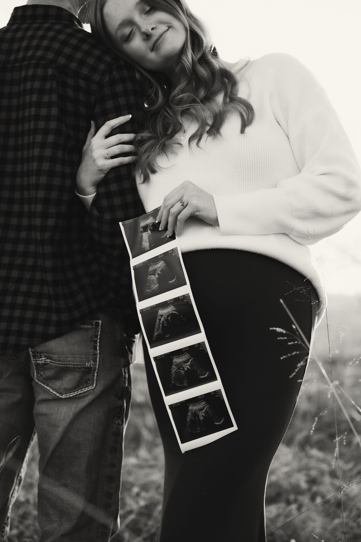 A black and white photo of a pregnant couple embracing while holding ultrasound photos in a field.