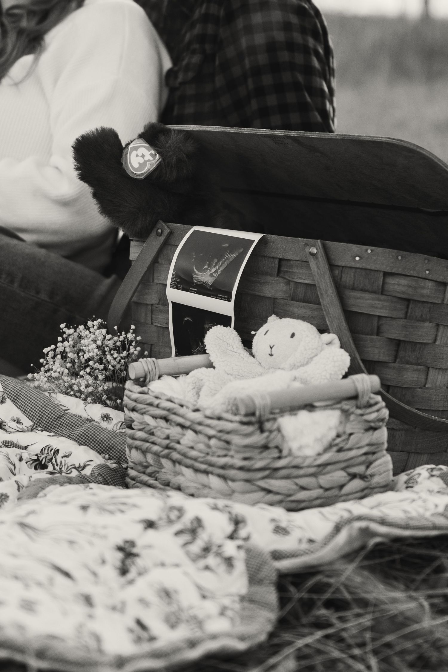 A black and white maternity photo of a couple sitting beside a wicker basket with baby items and ultrasound photos.