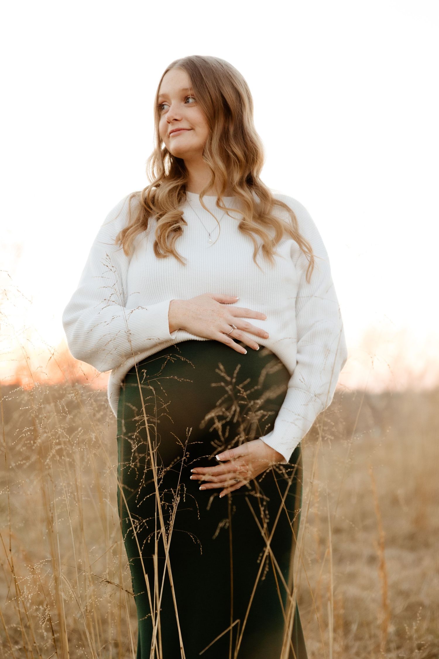 A glowing pregnant woman in a white sweater and green skirt cradles her bump in a golden sunlit field.